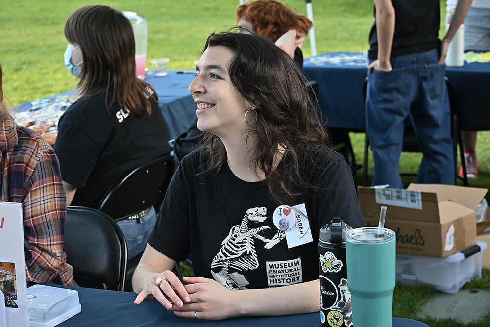 student tabling under tent on grass wearing shirt reading "museum of natural and cultural history" with dino skeleton and nametag reading "sarah"