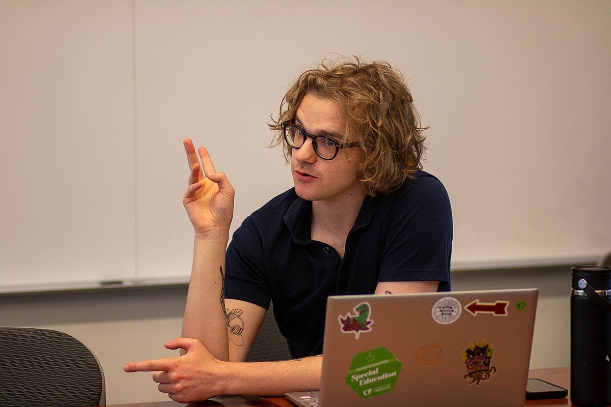 student speaking and gesturing seated at a meeting table with laptop