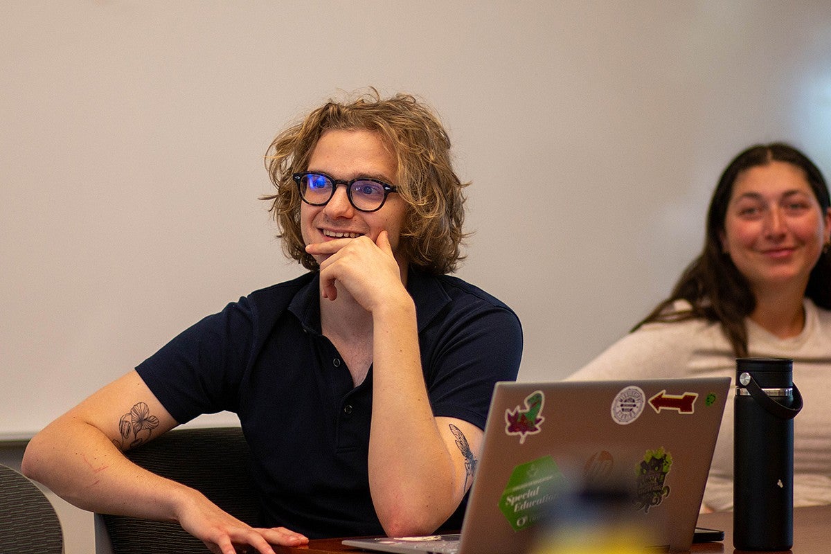 student seated at meeting table, listening to other students