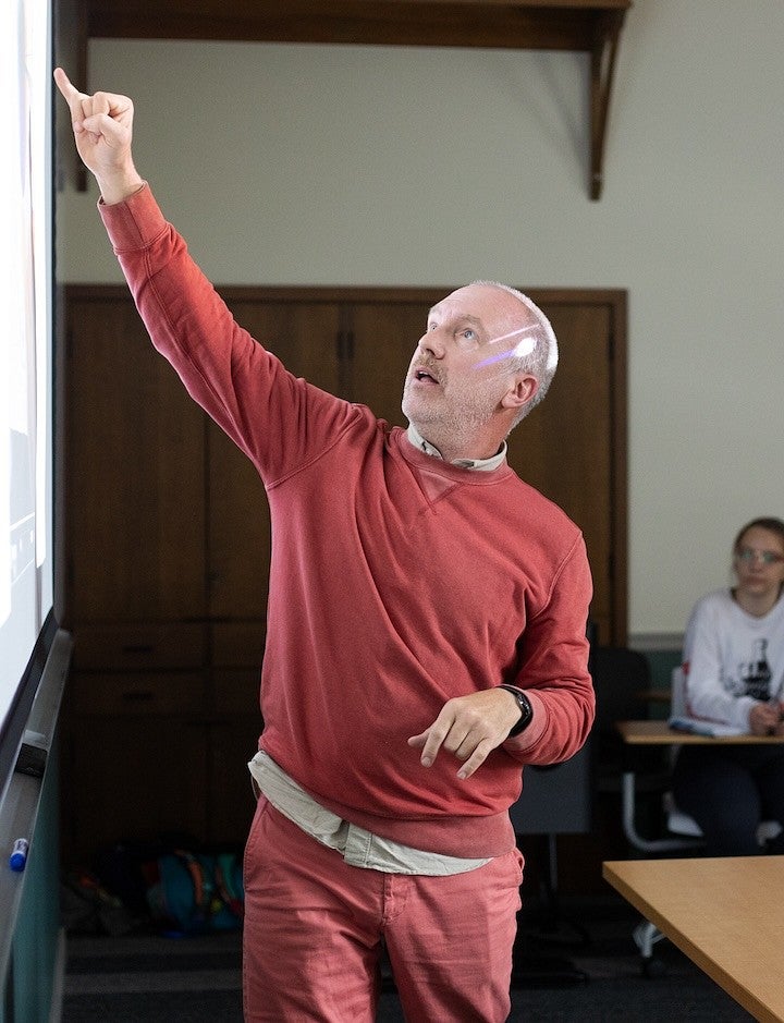 chris michlig standing at front of classroom, reaching up to point at the projector screen