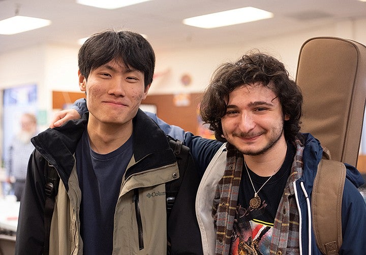 two students arm in arm, smiling at camera, one wearing guitar case