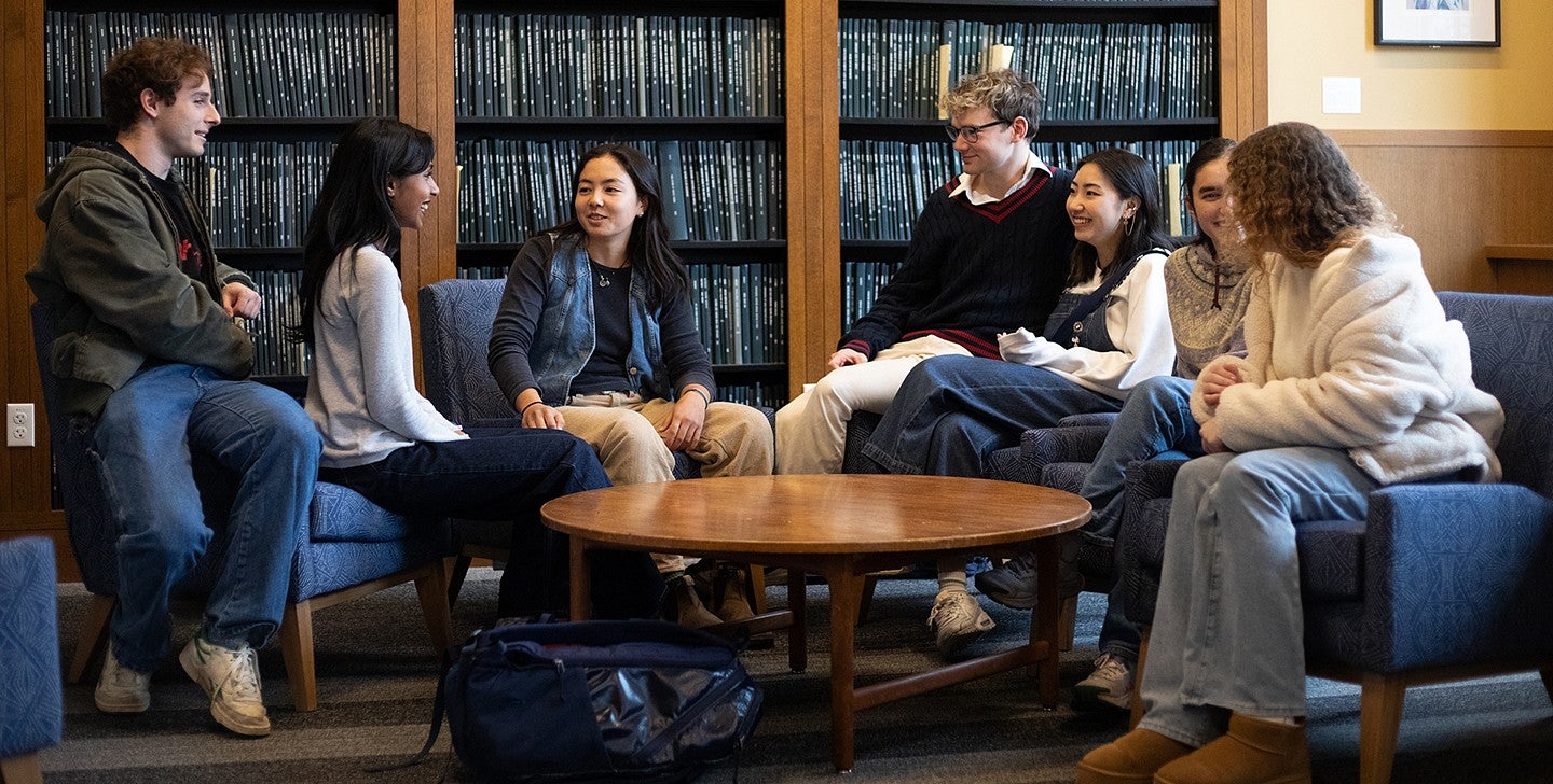 group of students seated in lounge chairs around low table in the Chapman Library in front of shelved theses, talking to one another