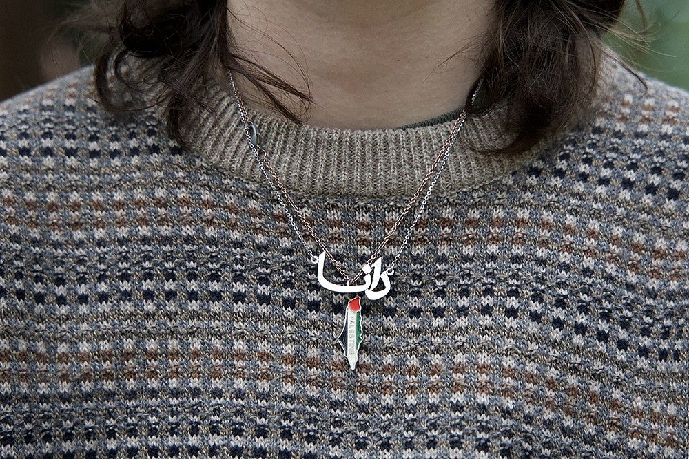 closeup of a necklace in the shape of gaza with arabic letters and its flag colors worn by a person in a sweater