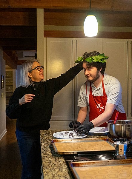 oliver loreto in his kitchen, baking, while his mother, Kate Mondloch, places a laurel wreath on his head