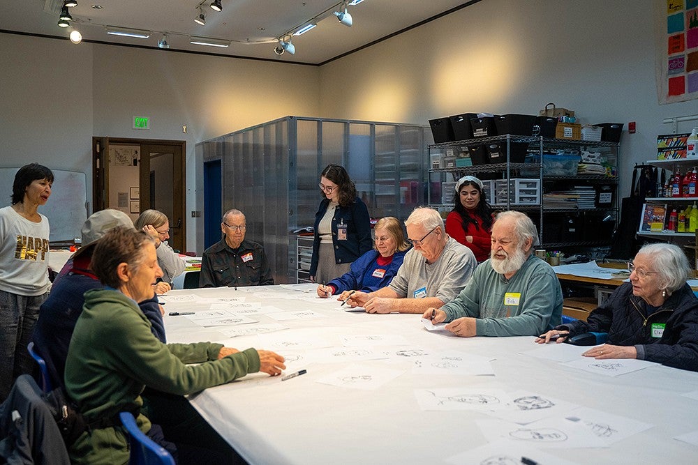 a table of senior adults working on drawings in an art studio, with younger teachers standing over them