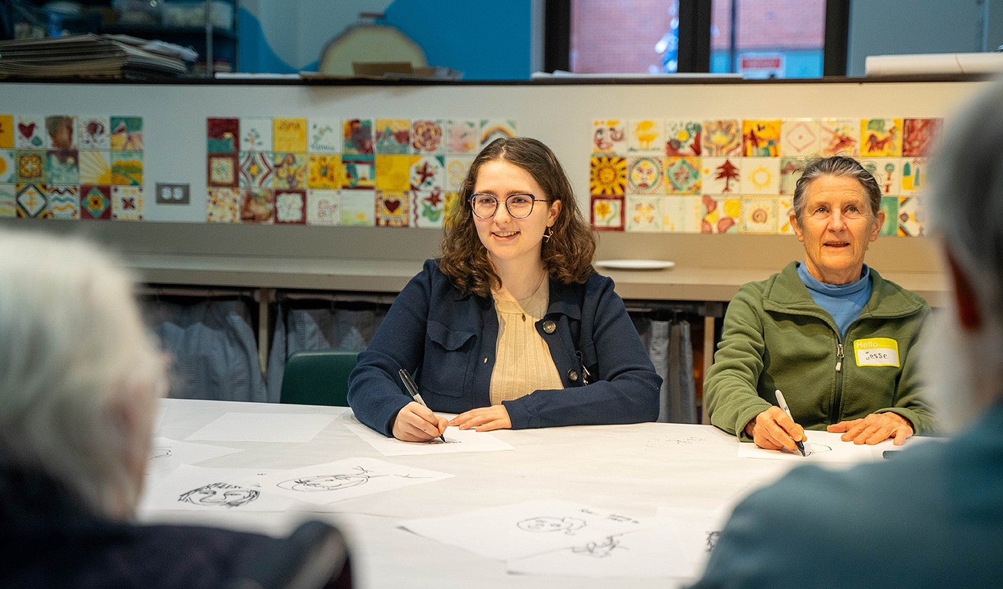 olivia black seated at a table in a classroom with senior-aged students and art decorating the walls