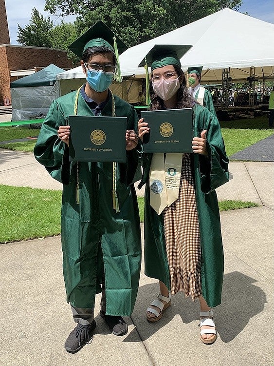 two students in grad regalia posing with diplomas outside and wearing masks