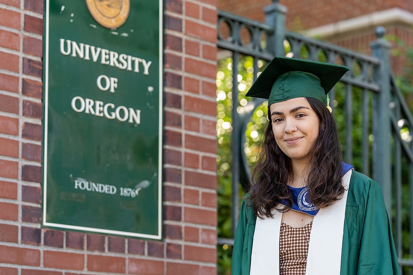 adrianna vaca-navarro wears a cap and gown and poses next to a UO campus sign