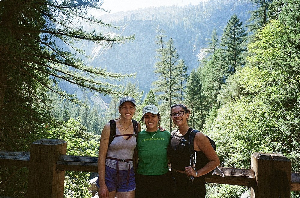 three young women posing in front of a wooden railing overlooking a forested view