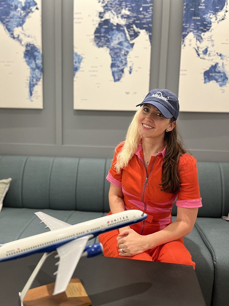 natalie jacobsen wearing an airlink cap seated at a table in front of a model plane, with world maps on the wall behind her