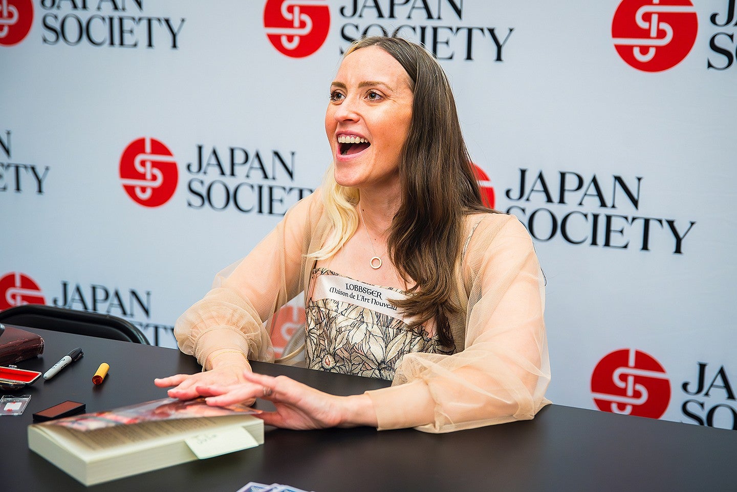 natalie jacobsen signing copies of her novel at a table with a wall of "japan society" logos behind her