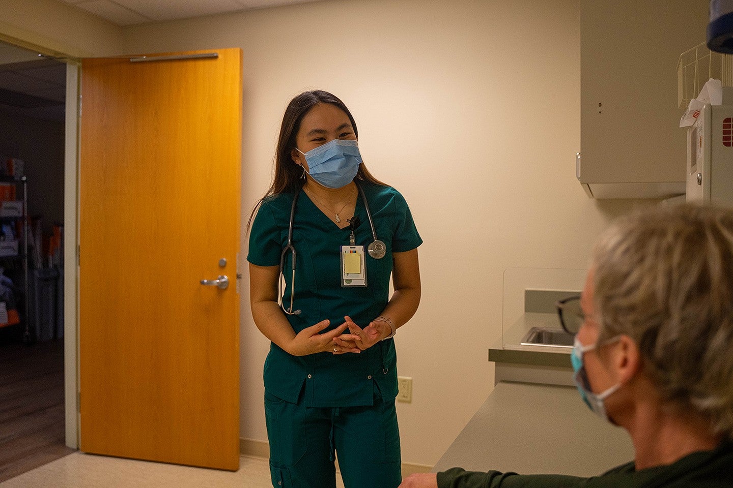 dora ho in medical scrubs and mask, talking to a patient in an exam room