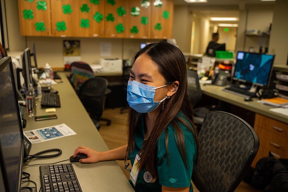 dora ho in scrubs and a mask, on a computer seated at a desk in a medical office