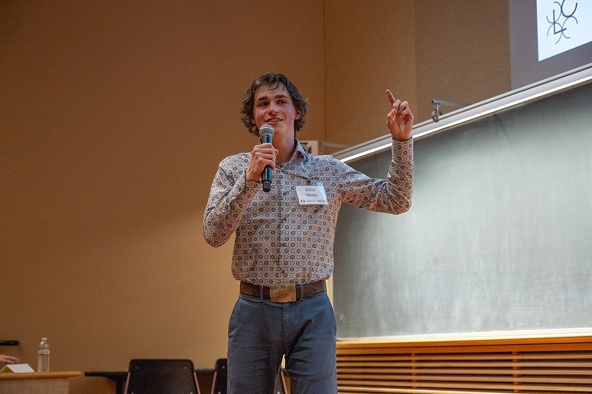 elliot nopp presenting at three minute thesis, gesturing with hand and holding mic
