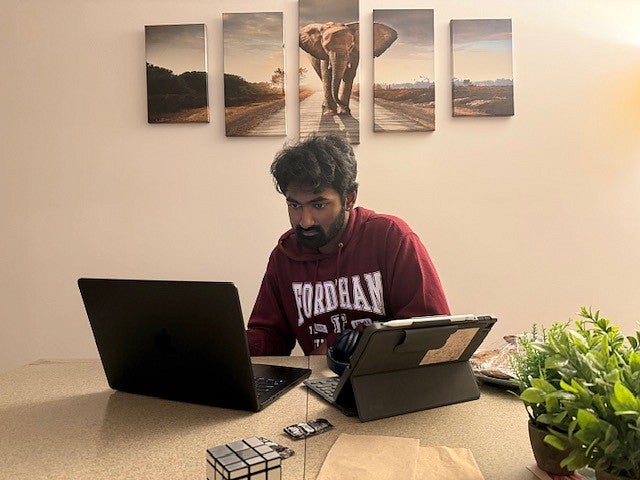 ashish sathya working on a laptop and a pad at a table in a fordham sweatshirt with elephant artwork behind him