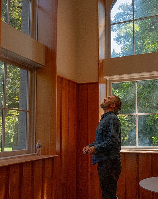 marcel brousseau standing at tall windows inside university hall, looking up at the trees