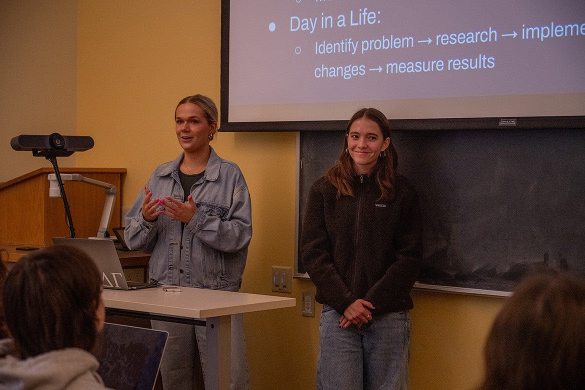 two students at front of a classroom, talking during a presentation with slides
