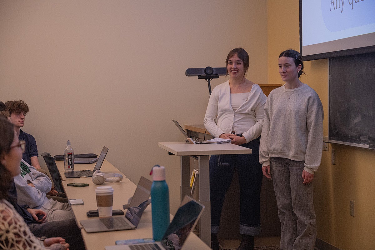 two students at front of a classroom, talking during a presentation with slides