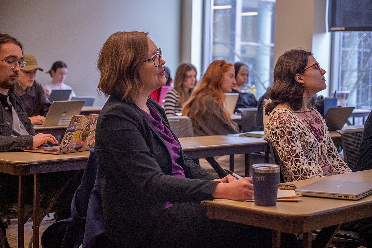 professor sitting among students in classroom, watching group presentations