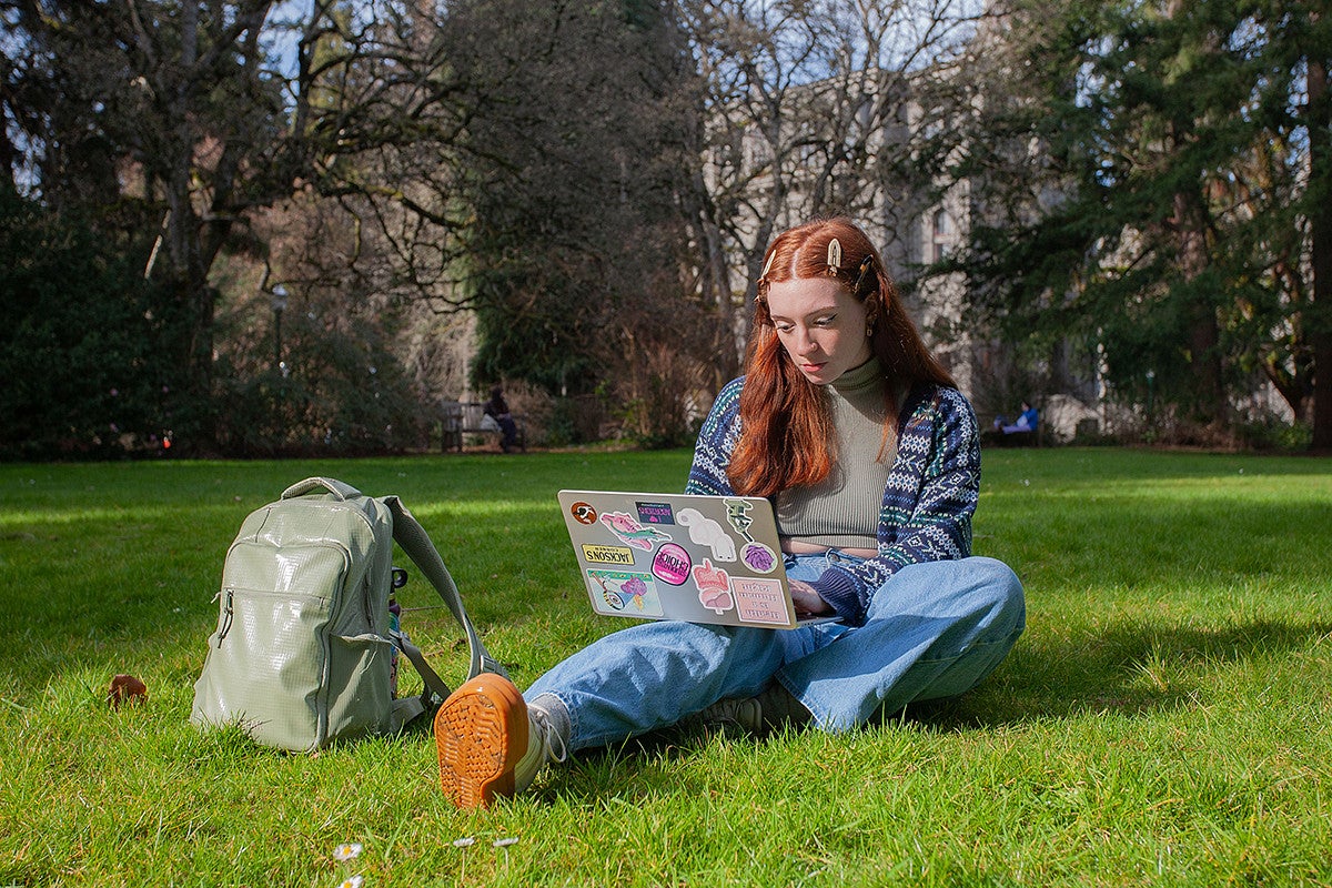 avery suriano seated with backpack and laptop on campus green, studying