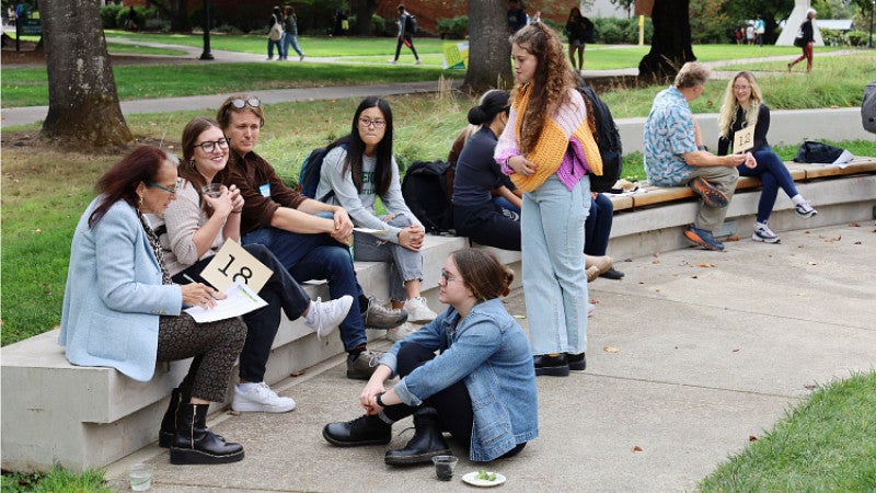 Professor Barbara Mossberg sits with a group of students on the edge of Tykeson-Chapman lawn