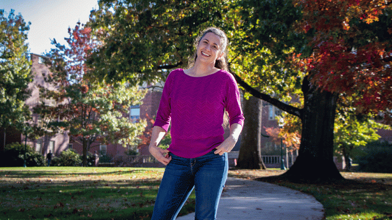 Lindsay Hinkle in a pink sweater standing under U of O campus oaks turning red in early autumn.