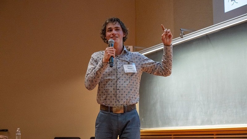 elliot nopp presenting at three minute thesis, gesturing with hand and holding mic