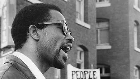 a black and white profile of a black man speaking in front of brick buildings.