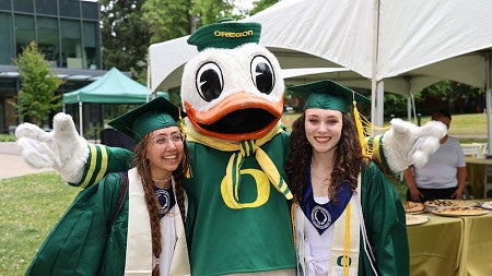 Two seniors in graduation robes are hugged by The Duck.