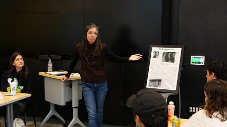 nicole dahmen teaching in a classroom, gesturing at a poster of a new york times front page