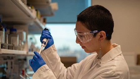 student waverly wilson in lab using pipette at bench, wearing blue gloves and white lab coat