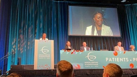 ann oluloro giving a speech at a conference, standing at podium on stage with herself projected on screen behind her