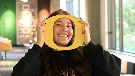 student in campus building holding yellow cutout O around their face, smiling