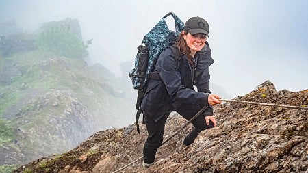 person in backpack and Oregon cap climbing up rock face on misty ridge, smiling at camera