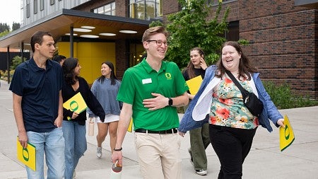 student leading campus tour, laughing with tour participants holding yellow folders and walking past a building
