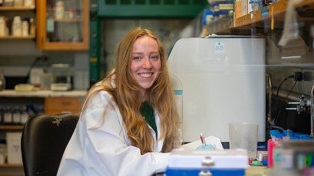 student in lab coat seated at lab bench, holding pen and smiling over her shoulder