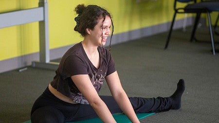 student in yoga attire sitting on mat with legs straddled, leaning forward and smiling