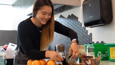 student working with fruit and coffee grounds next to laptop in campus kitchen