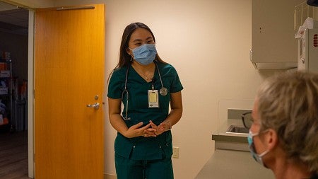 dora ho in medical scrubs and mask, talking to a patient in an exam room