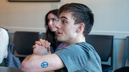 student sitting at table with others, talking animatedly, with peace sign tattoo above elbow