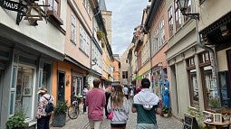 a european streetscape with cobblestones and shops, following three students walking down the middle of the street