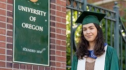 adrianna vaca-navarro wears a cap and gown and poses next to a UO campus sign