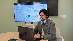 man sitting at laptop at conference table with graphs on screen behind him