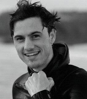 Black and white head shot of Aaron Georis in a wetsuit, blurred body of water in background.