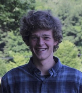 Head shot of Sam Schroeder wearing plaid and standing in front of trees.