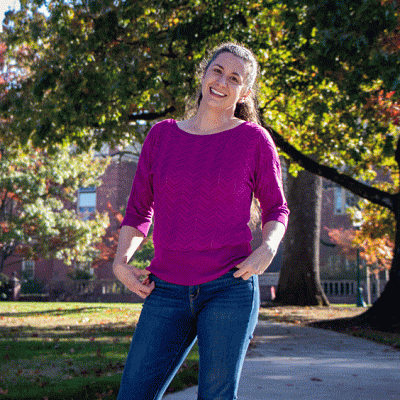 Lindsay Hinkle in a pink sweater standing under U of O campus oaks turning red in early autumn.