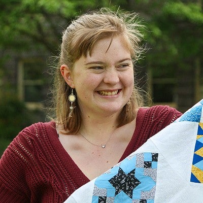 portrait of Trish Young outdoors, smiling off camera, holding up colorful quilt