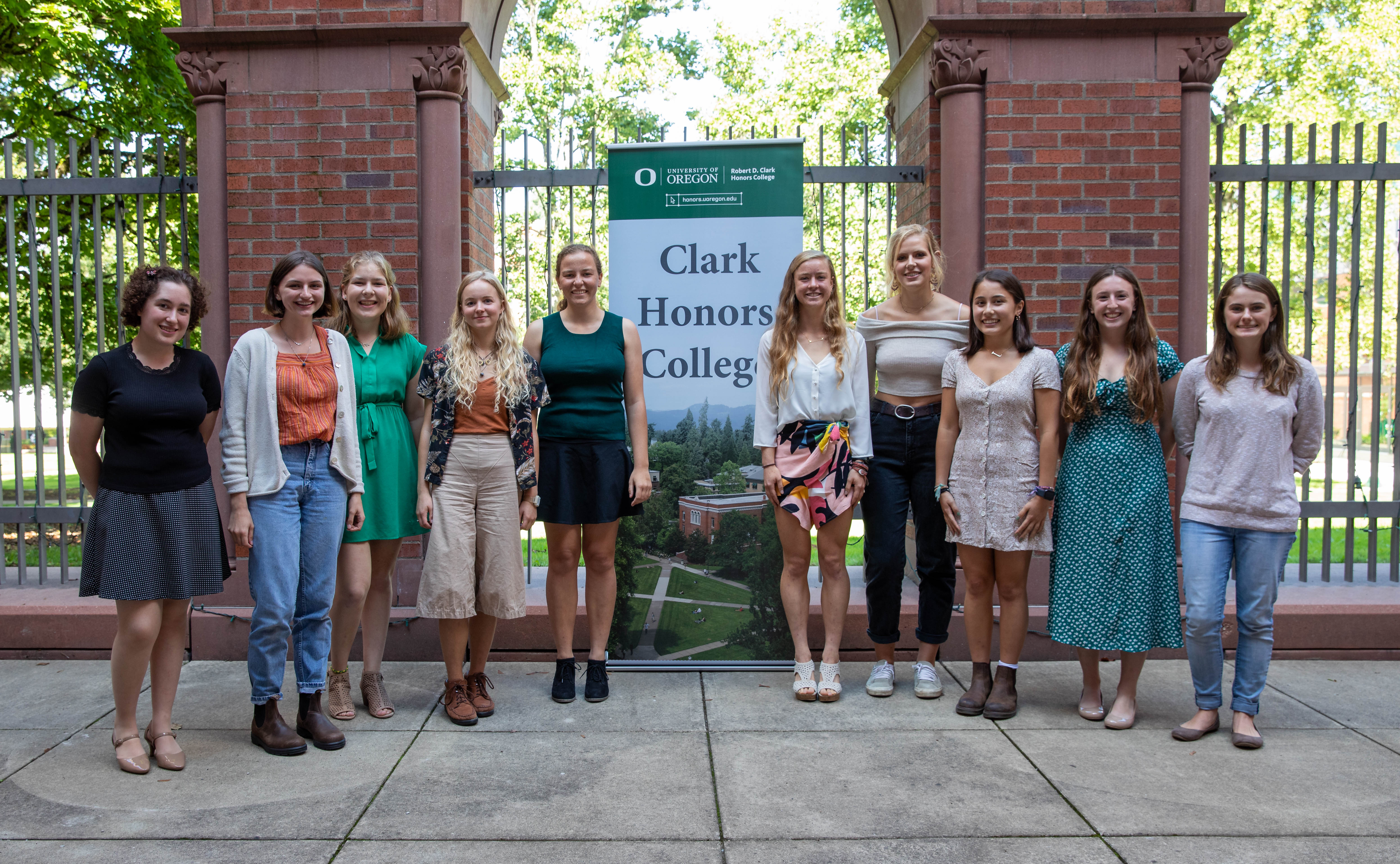 A group of students standing in front of a wrought iron fence of an outdoor courtyard in front of a sign reading &quot;Clark Honors College.&quot; 