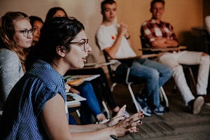 A woman leans forward in her chair while speaking to a circle of seated students.