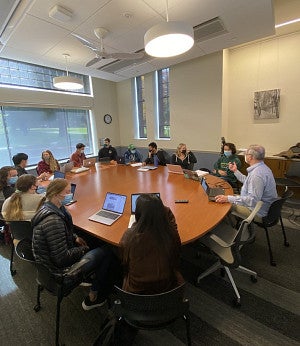 Students and a male professor, masked against COVID, with laptops, seated around a classroom table.